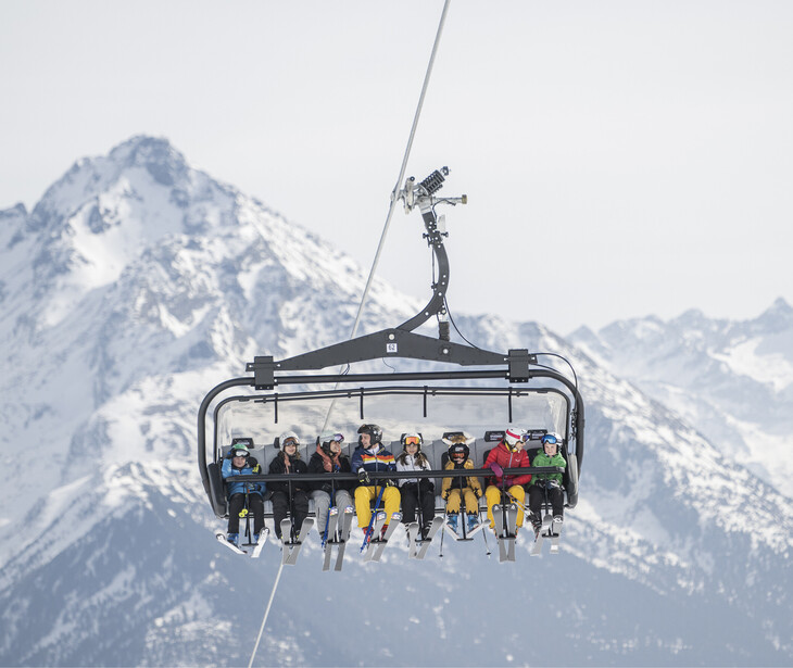 Sesselbahn mit mehreren Skifahrern vor schneebedecktem Bergpanorama.