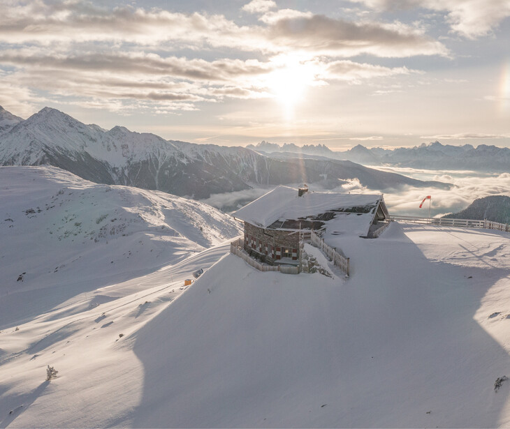 Berghütte auf schneebedecktem Hang mit Bergpanorama und Sonnenuntergang im Hintergrund.