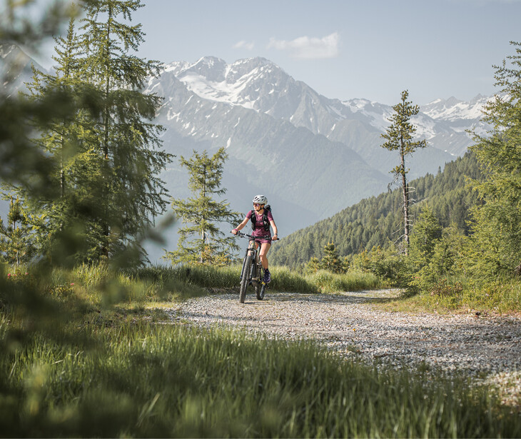 Frau mit Helm und Rucksack fährt mit dem Fahrrad auf einem Schotterweg mit Bergpanorama im Hintergrund.
