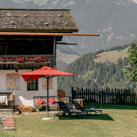 Chalet mit Holzschindeldach, Balkon mit roten Blumen, rotem Sonnenschirm und Liegestühlen vor Bergkulisse.