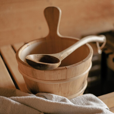 Holzeimer mit Holzlöffel auf Holztisch neben grauem Handtuch in Sauna.