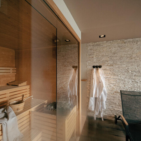 Saunabereich mit Holzbänken hinter Glas, zwei weißen Bademänteln an der Wand und einem schwarzen Stuhl auf dunklem Boden.