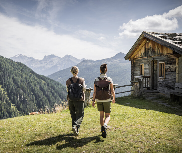 Zwei Wanderer mit Rucksäcken gehen auf einer Wiese an einer Holzhütte vorbei in Richtung Bergpanorama.