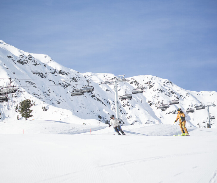 Skifahrer auf schneebedeckter Piste vor Berglandschaft mit Sessellift im Hintergrund.