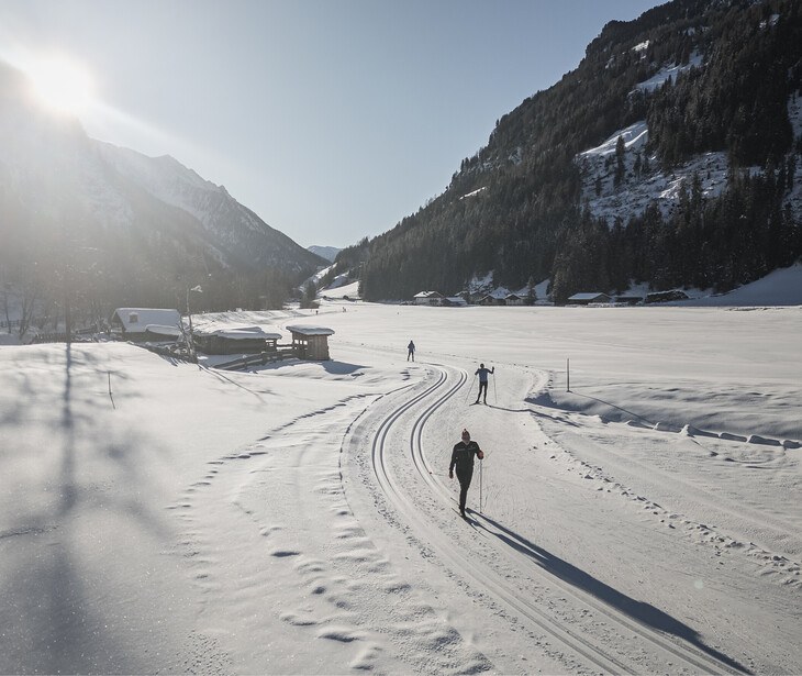 Drei Langläufer auf einer gespurten Loipe in einem schneebedeckten Tal mit Bergen im Hintergrund bei Sonnenschein.