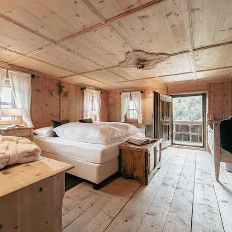 Rustikales Schlafzimmer mit Holzbalkendecke, Doppelbett, Holzmöbeln und offenem Fenster mit Blick auf einen Balkon.