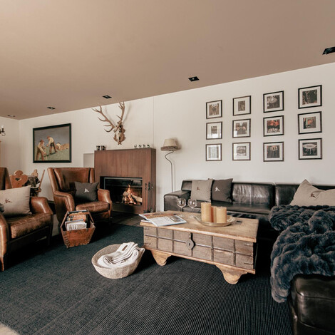 Wohnzimmer mit Ledersesseln, Ledercouch, Kamin, Holztisch und Wanddekoration mit Geweih und gerahmten Bildern.