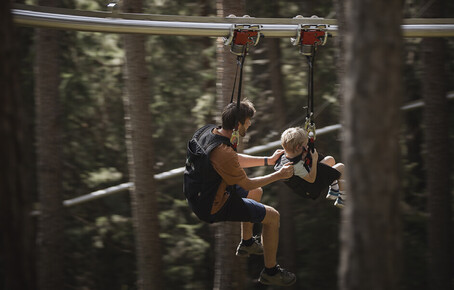 Zwei Personen sitzen hintereinander an einem Zip-Line duch einen Wald.