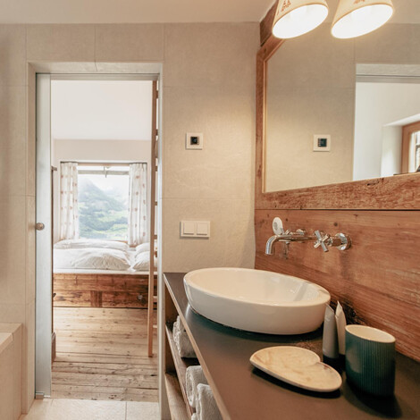 Badezimmer mit Badewanne, Handtuchheizkörper und Waschbecken mit Holzspiegel, Blick durch Tür in Schlafzimmer mit Bett und Fenster.
