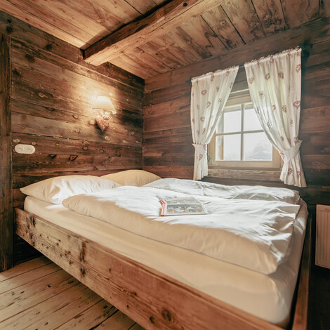Rustikales Schlafzimmer mit Holzwänden, Holzboden, Doppelbett mit weißen Bettwaren, Fenster mit hellen Vorhängen.