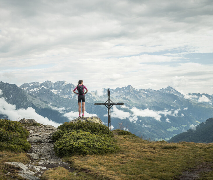 Wanderin steht auf einem Berggipfel neben einem Gipfelkreuz mit Blick auf eine Bergkette unter bewölktem Himmel.