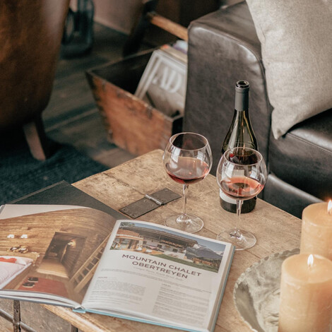 Wohnzimmertisch mit aufgeschlagenem Magazin, zwei Weingläsern, brennenden Kerzen und Ledersofa mit Kissen im Hintergrund.