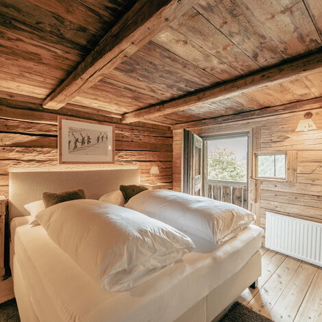 Schlafzimmer mit Holzwänden und -decke, Doppelbett mit weißen Bettdecken, Nachttisch mit Lampe und offenem Fenster mit Blick ins Grüne.