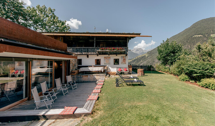 Chalet mit Terrasse, Liegestühlen und Garten vor Bergkulisse unter blauem Himmel.
