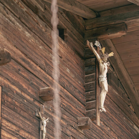 Kruzifix an der Außenwand eines Holzhauses unter einem Dachvorsprung.