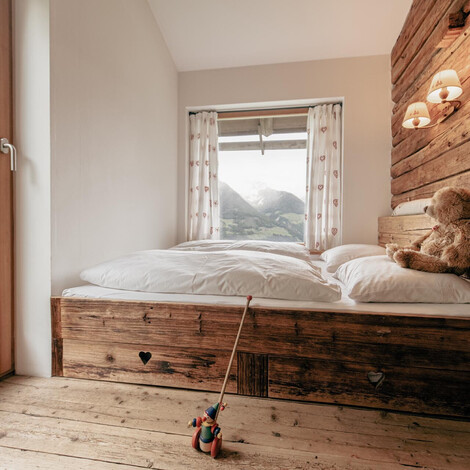Schlafzimmer mit Holzbett, Teddybär auf dem Kopfkissen und Blick auf Berge durch Fenster mit Vorhängen.