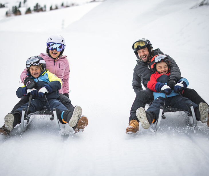 Zwei Erwachsene und zwei Kinder sitzen auf Schlitten und fahren einen schneebedeckten Hang hinunter.
