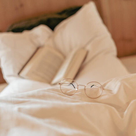 Brille mit rundem Gestell liegt auf weißer Bettdecke vor aufgeschlagenem Buch und Kopfkissen.