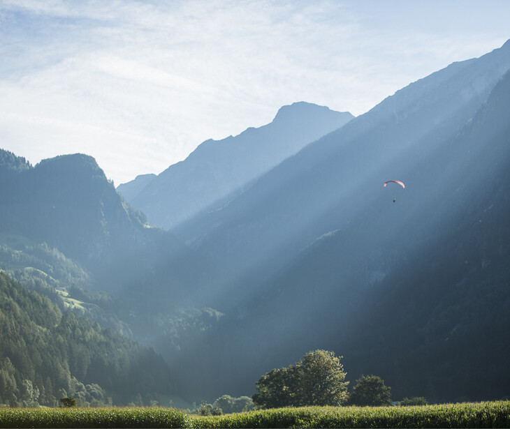 Berglandschaft mit bewaldeten Hängen, Talwiese und einem Gleitschirmflieger am Himmel.