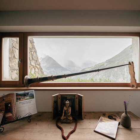 Langes Alphorn liegt auf einem Fensterbrett mit Bergblick, darunter stehen Bücher, eine Holzfigur und eine Vase mit Blume auf einem Holztisch.