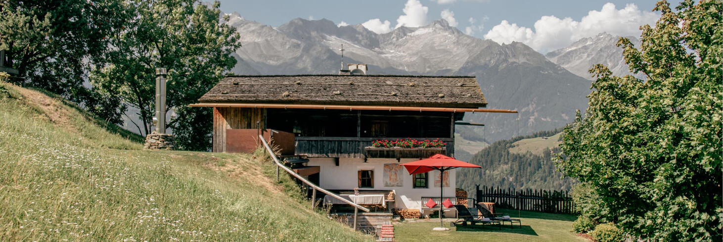 Chalet auf einer Wiese mit Bäumen und Bergen im Hintergrund unter blauem Himmel mit Wolken.